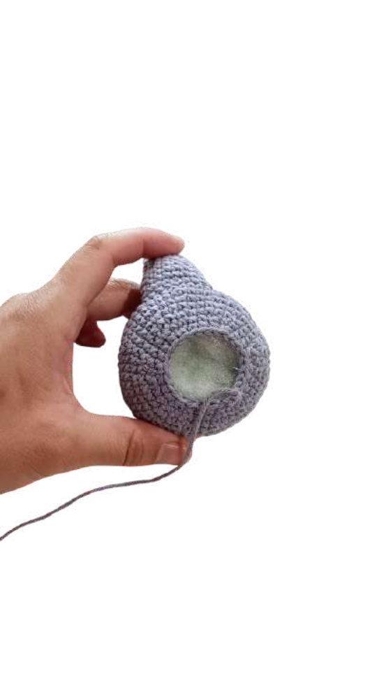 A hand holds a partially completed grey crochet sphere for an amigurumi project, with an opening revealing the stuffing inside.