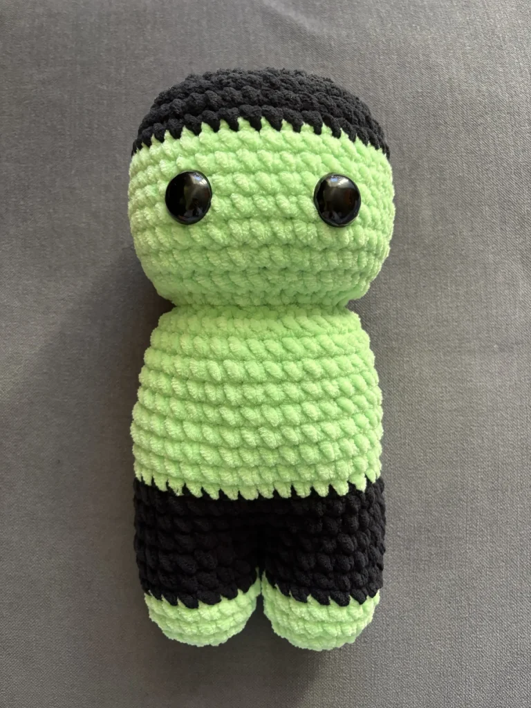 Crocheted Frankenstein plushie with a light green head and torso, black hair, and black legs, featuring two large black plastic eyes.