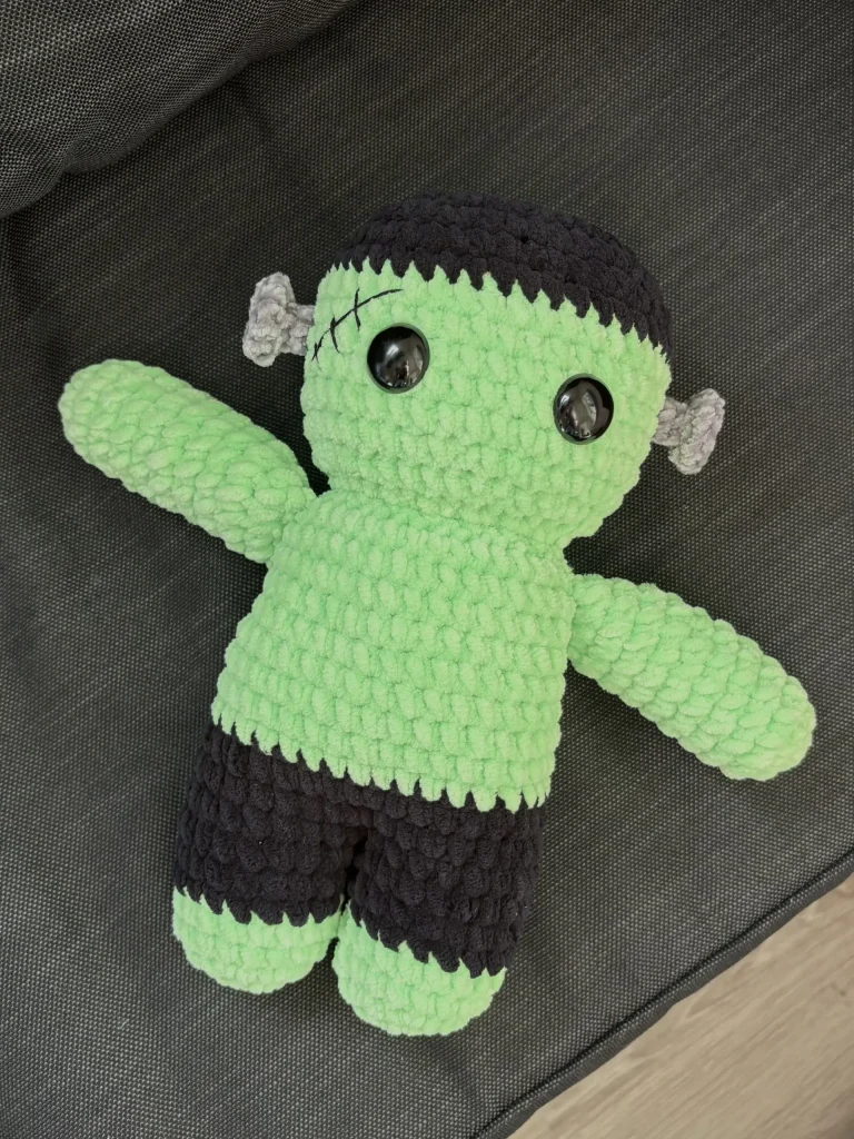 Crochet plushie of Frankenstein’s monster with green skin, black hair, and gray neck bolts.