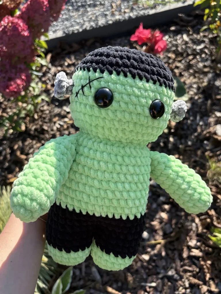 A crocheted Frankenstein plushie with green skin, black hair, and black pants.