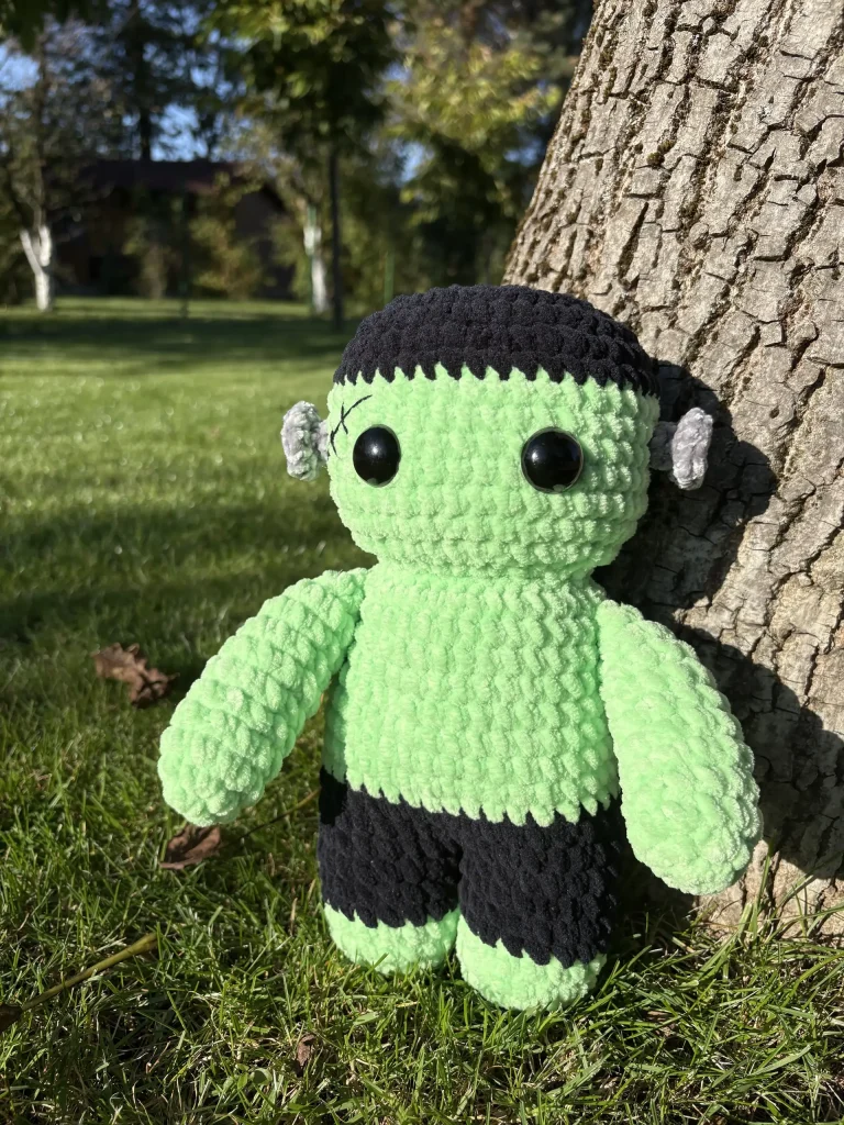 A Frankenstein plushie with black hair, a stitched forehead, and green skin stands in the grass against a tree trunk, perfect for a Halloween crochet pattern display.