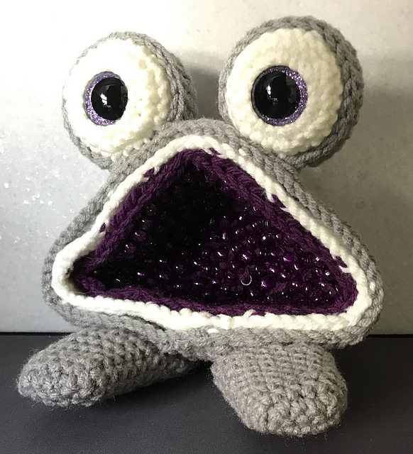 Grey crocheted monster with large eyes and a wide, open mouth filled with purple beaded crystals, crafted using crochet geode patterns.