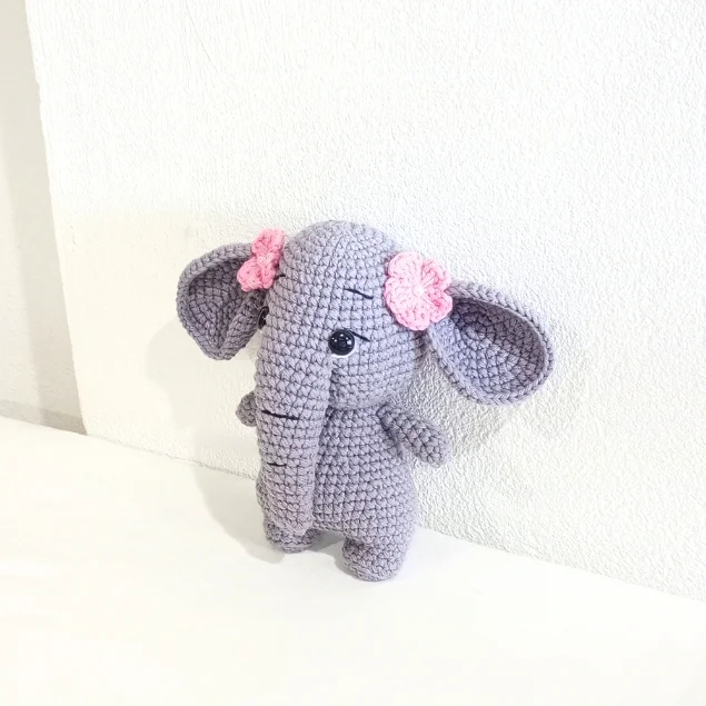 A gray crochet elephant toy adorned with two pink flower hair clips, crafted using a crochet pumpkin pattern style.