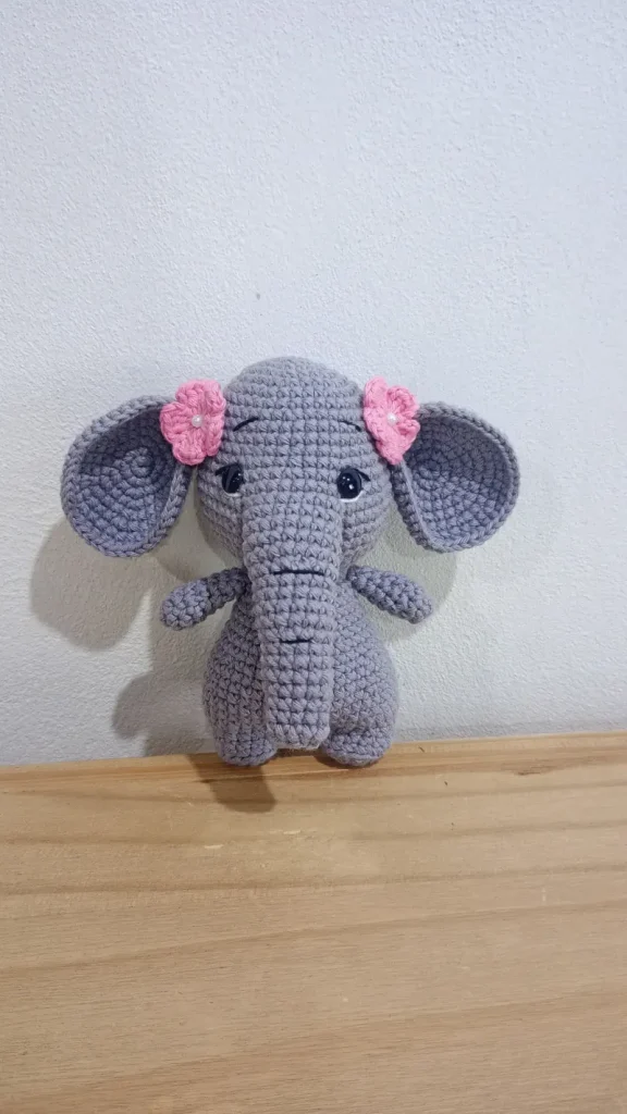 Crocheted gray elephant amigurumi toy with small pink flowers on its ears, created using an elephant amigurumi crochet pattern.