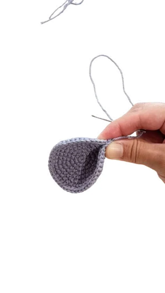 A hand holding a partially crocheted light blue circular piece for an elephant amigurumi project, with a loose thread attached.