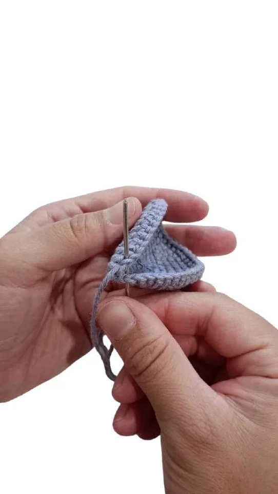 Hands using a needle to sew two pieces of light blue crocheted fabric for an elephant amigurumi together.