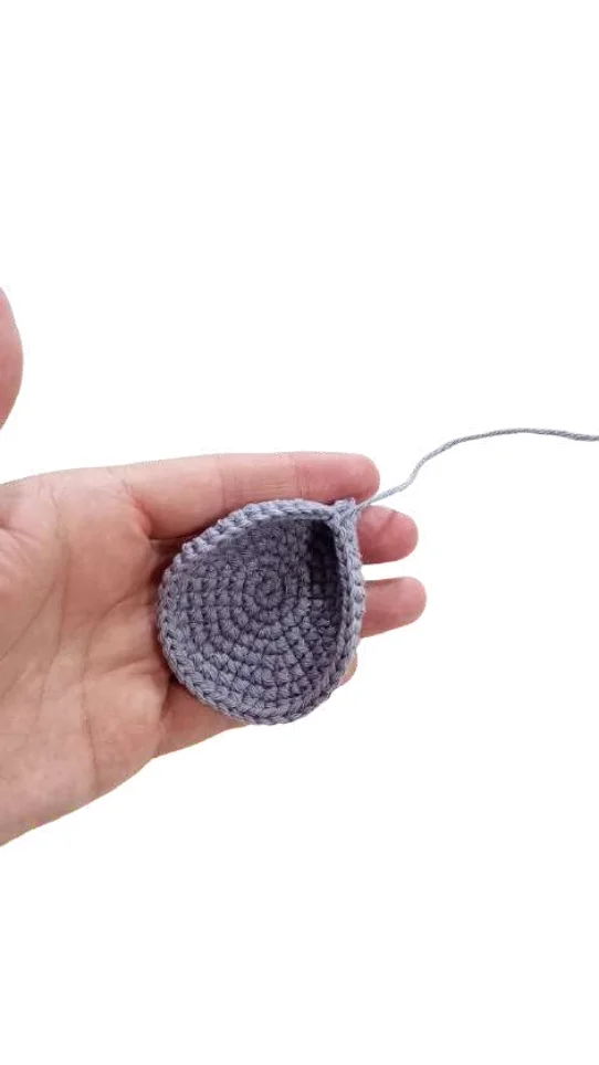 A hand holds a small, circular piece of grey crochet work for a pumpkin crochet pattern, with a yarn tail attached.