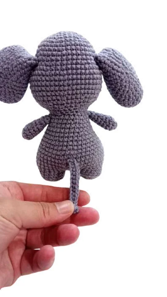 A hand holds a small, gray elephant amigurumi by its tail.