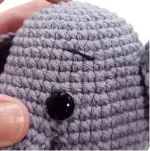 Close-up of a grey elephant amigurumi held in a hand, featuring a black plastic eye and an embroidered eyebrow.