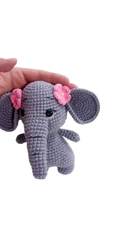 A small, grey crocheted elephant toy with pink flower accents on its head, held in a hand alongside a crochet pumpkin pattern.