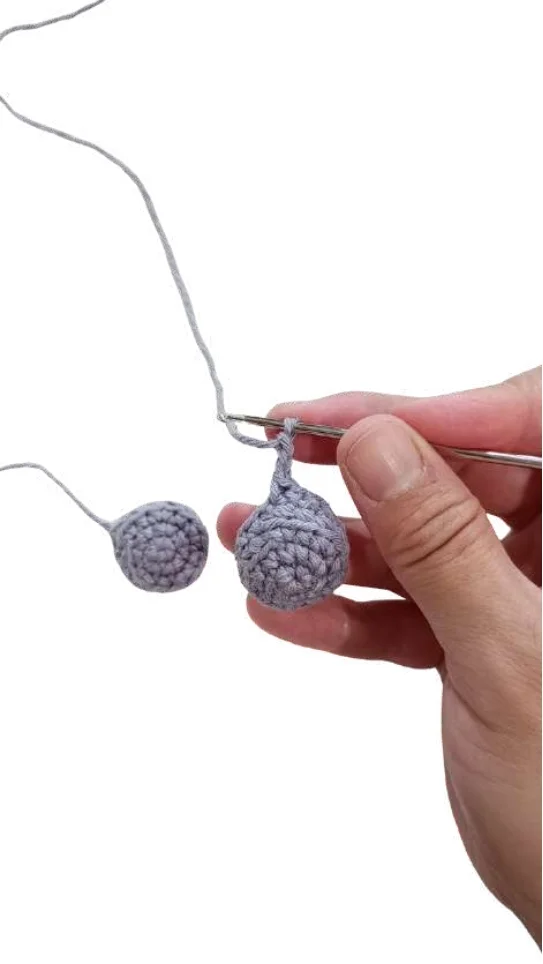 A hand holding a crochet hook, attaching a gray crocheted sphere to another to create an elephant amigurumi with gray yarn.