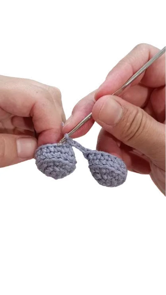Hands using a metal hook to crochet two joined gray pieces for an elephant amigurumi.