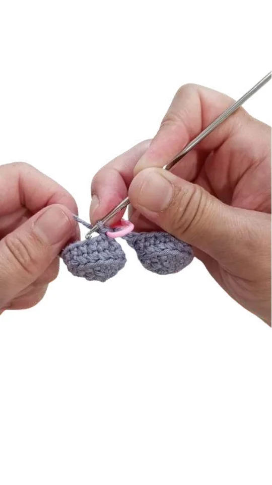 Hands use a metal crochet hook to join two grey pieces for an elephant amigurumi, secured with a pink stitch marker.