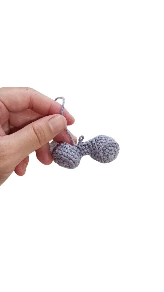 A hand holds a small, gray piece shaped like two connected spheres, a simple crochet pumpkin pattern.