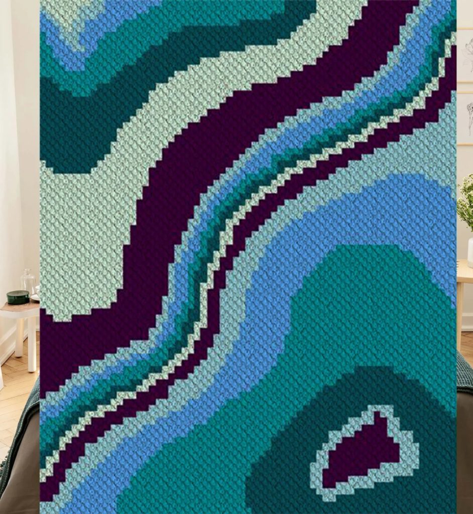 A knitted blanket featuring an abstract, pixelated pattern of wavy bands in shades of blue, teal, and dark purple, inspired by crochet geode patterns.