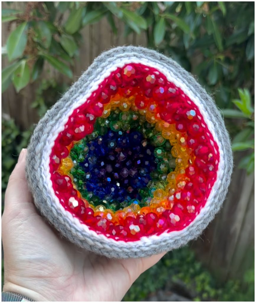 A geode-shaped object made using crochet geode patterns, featuring a grey exterior and a rainbow-colored interior lined with faceted beads.