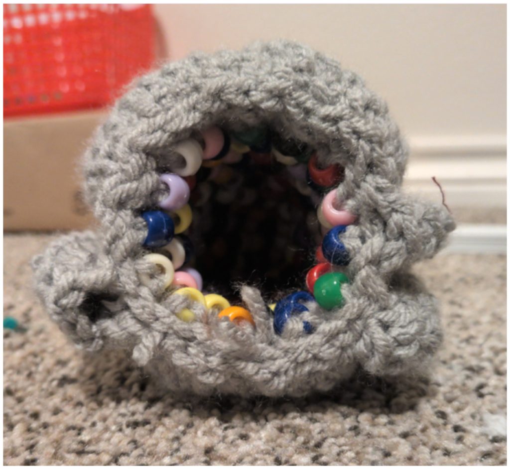 A grey crochet geode pattern tube lined with colorful plastic pony beads along the opening.