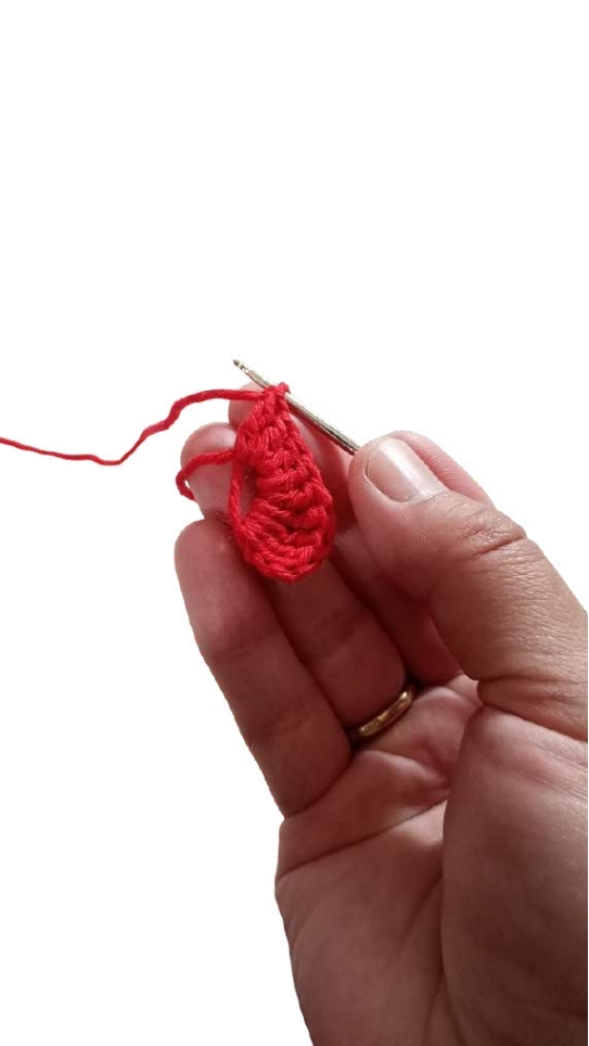 A hand holds a crochet hook while working on a small red circular piece for a crochet pumpkin pattern.