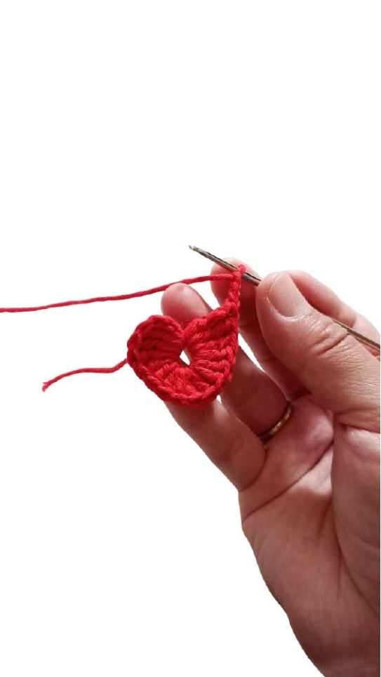 A hand holds a crochet hook, working on a red yarn heart-shaped piece that could be a gift for a giraffe amigurumi.