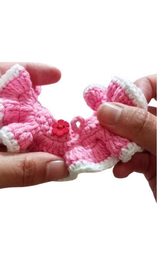 Hands hold a small, pink and white crocheted accessory with a red flower button, similar in scale to a giraffe amigurumi.