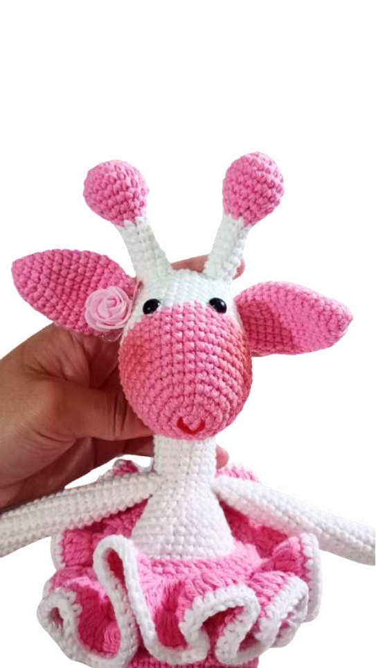 A hand holds a white and pink giraffe amigurumi wearing a pink tutu and a small flower on its ear.