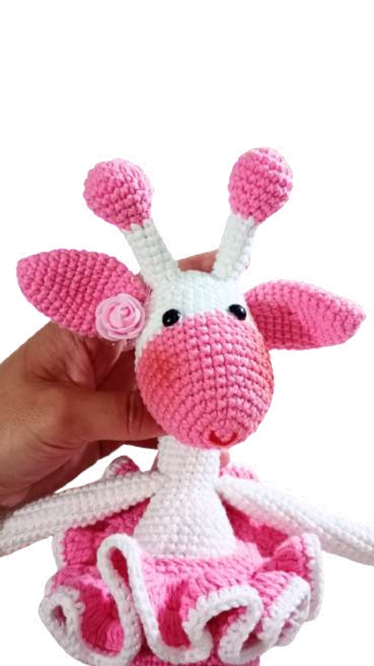 A hand holds a crocheted giraffe amigurumi toy with a white head, pink snout, pink ears, and a pink tutu.
