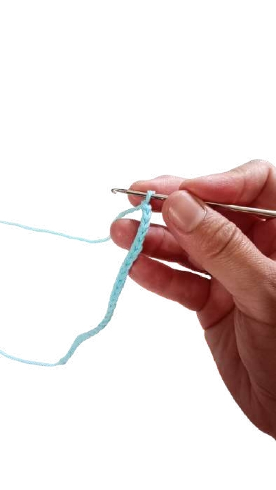 A hand holding a crochet hook, working a chain of light blue yarn for a giraffe amigurumi.