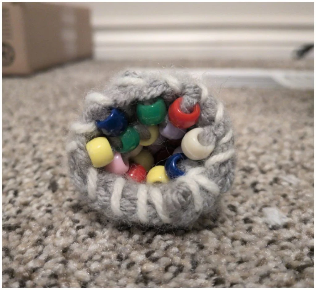 A small, grey hand-knit pouch containing assorted colorful plastic beads, reminiscent of crochet geode patterns.