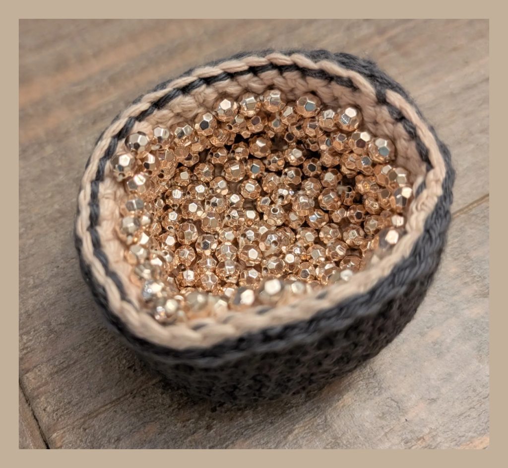 Small, round, crocheted bowl in a geode pattern, filled with metallic, faceted gold beads.